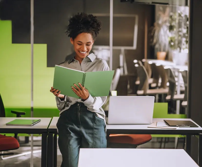 Smiling woman reading green notebook in modern office workspace with laptop