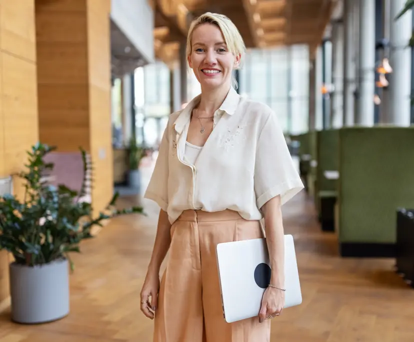 Smiling blonde businesswoman in beige trousers holding laptop in open-plan modern office