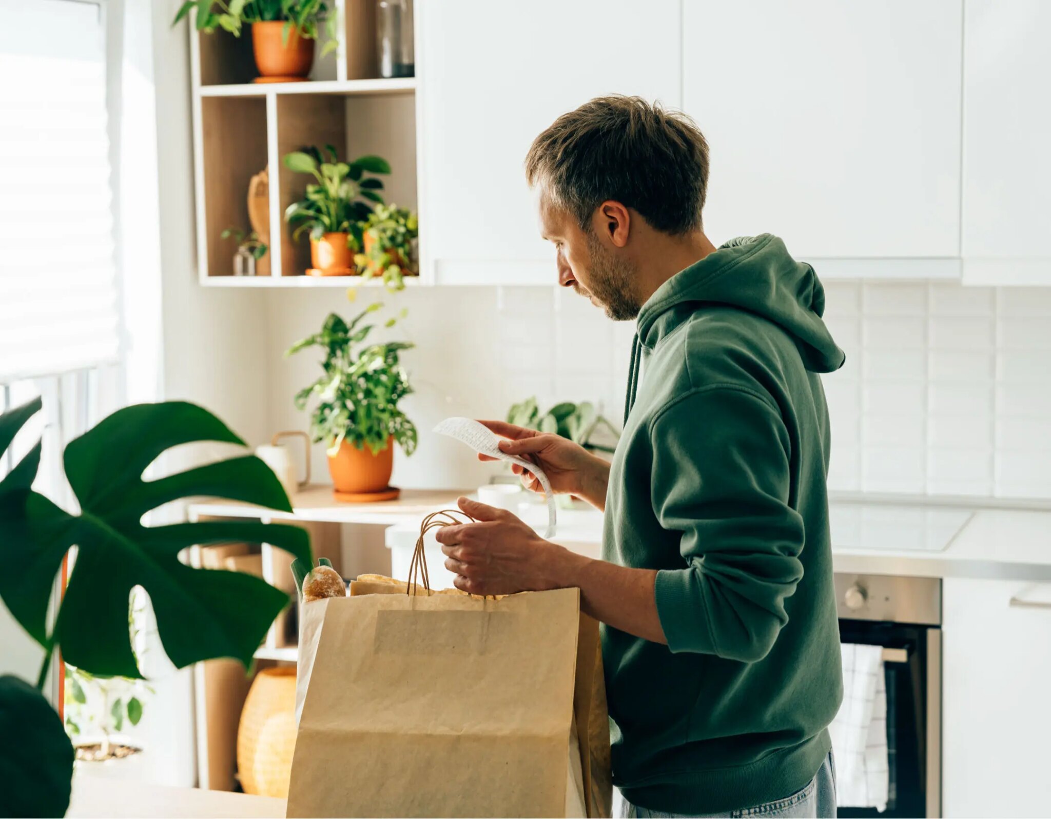Man in green hoodie reviews grocery receipt while unpacking bag in plant-filled kitchen.