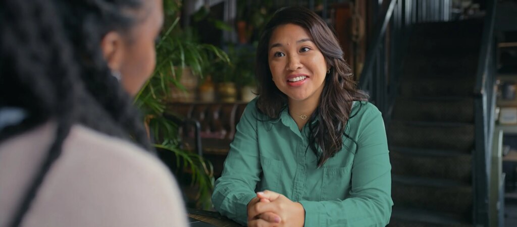 Smiling Asian mortgage broker in green shirt consulting client at plant-filled cafe office