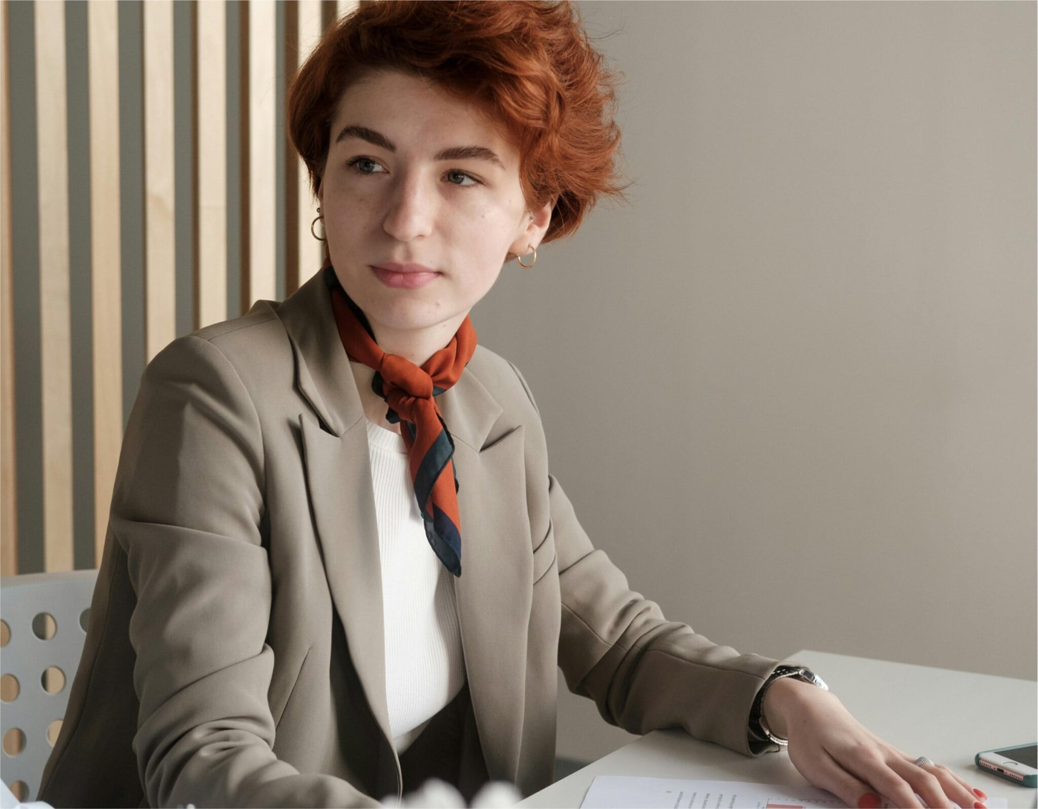 Red-haired professional woman in beige blazer and orange scarf at modern office desk