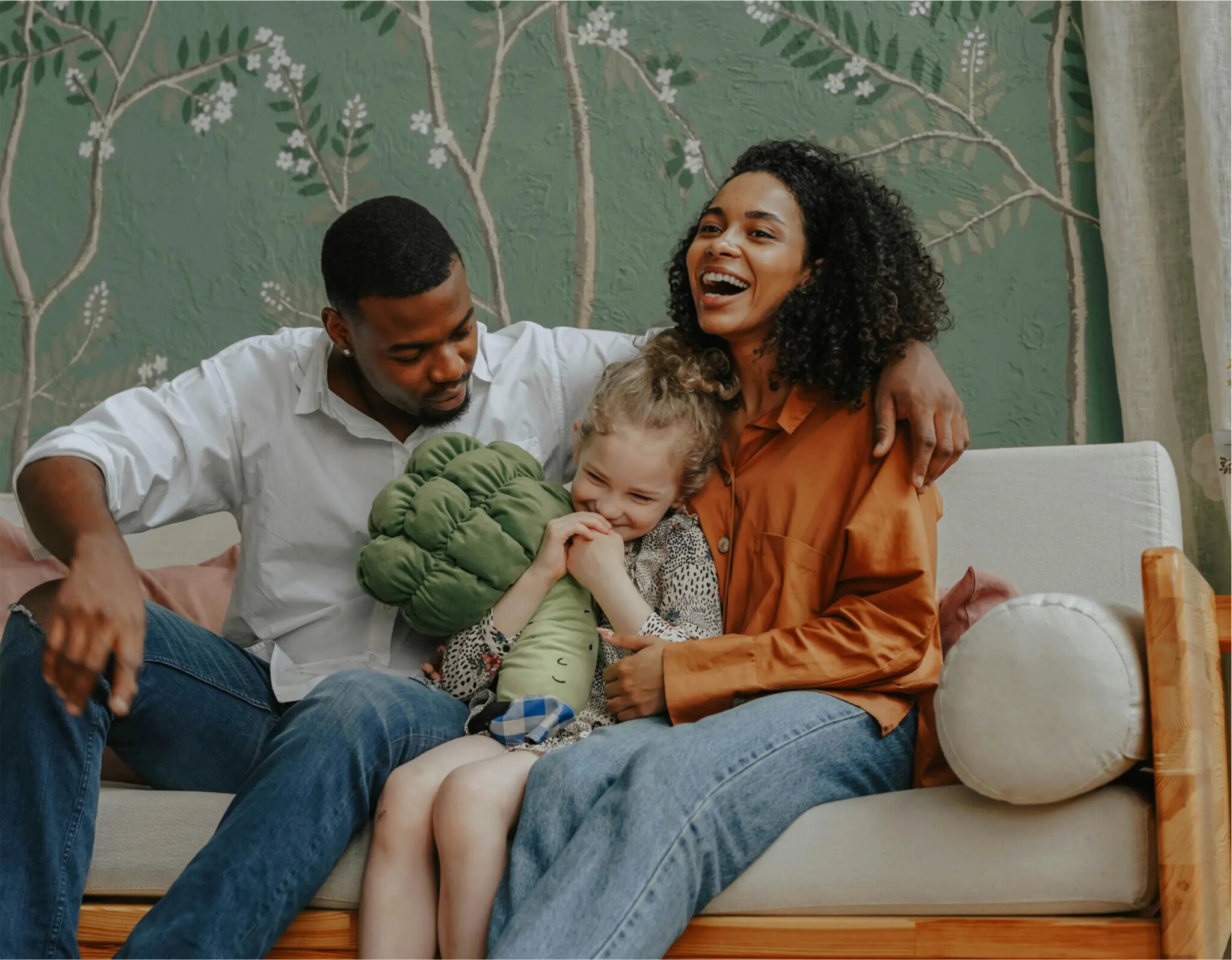Black family of three on beige sofa with green floral wall holding plush toy