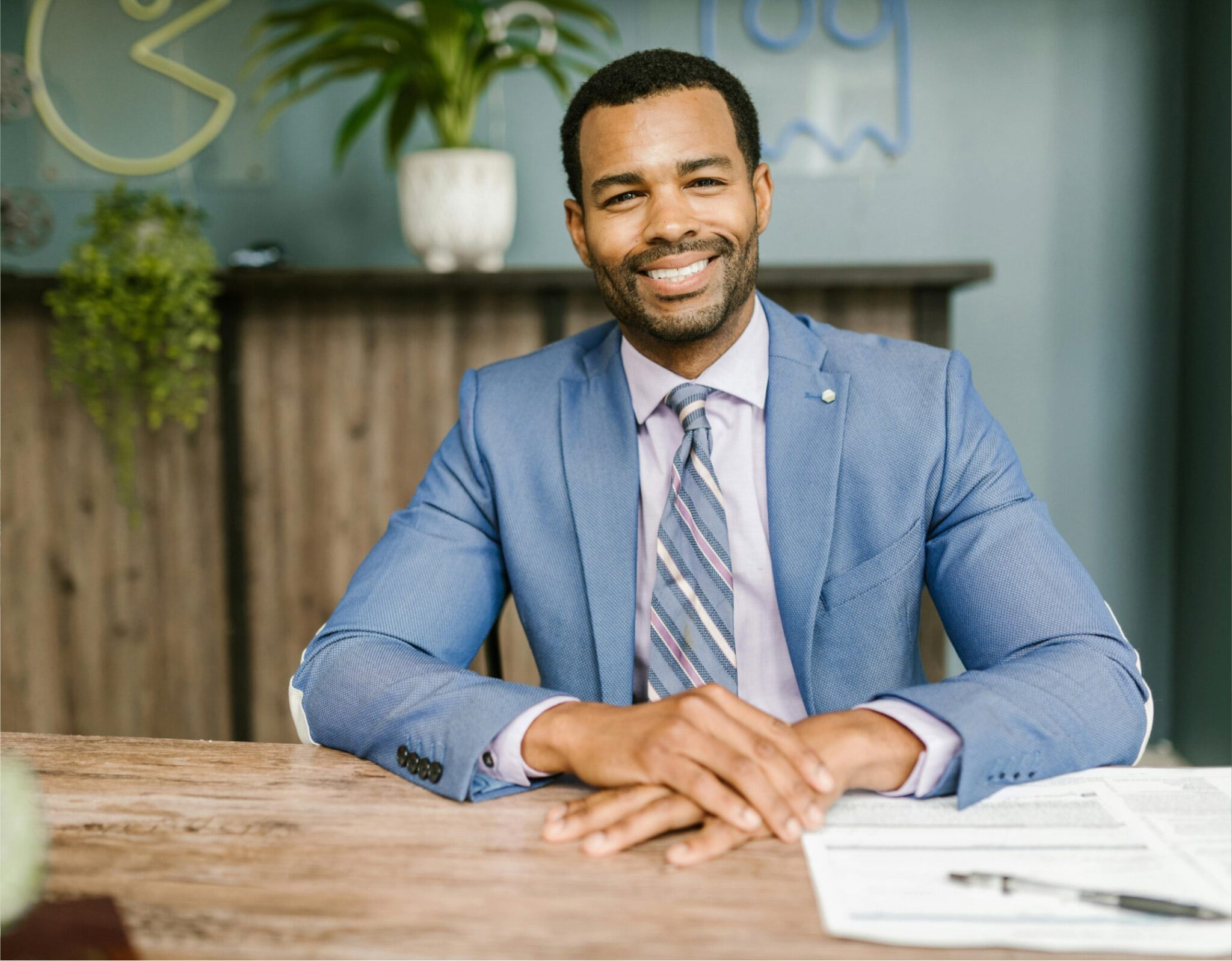 Smiling Black financial advisor in blue suit and striped tie at wooden desk with potted plant