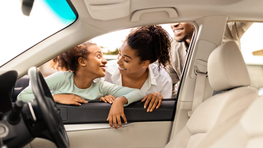 Mother, daughter and father smiling through open car door in sunny driveway